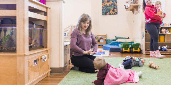 A woman sits on the floor showing a card to a baby in a pink dress, while another woman holds a child and stands in the background of a classroom.