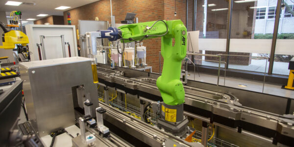 A green robotic arm operates on an automated assembly line in a modern industrial facility with glass windows and brick walls in the background.