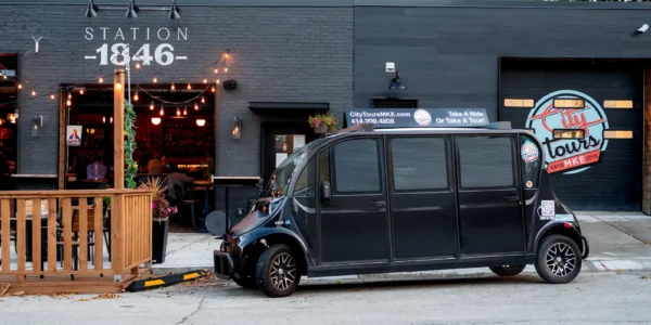 A black tour vehicle is parked in front of a dark gray building labeled “Station 1846” with outdoor seating and decorative string lights.