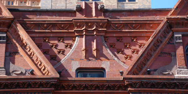 Detailed red brick and stone building facade with decorative carvings, triangular roof sections, and a small window centered below ornate patterns.