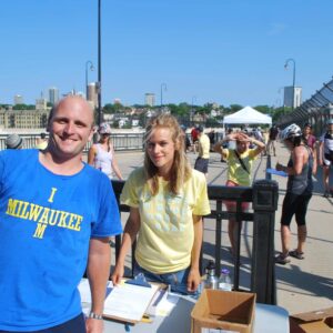 Man in “I Milwaukee” shirt and woman with clipboard stand at an outdoor registration table while people gather and interact on a sunny day.