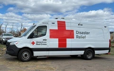 A white American Red Cross van with a large red cross and “Disaster Relief” text is parked outdoors on a cloudy day.