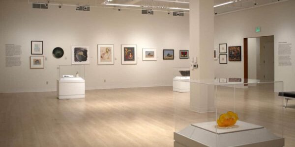A modern art gallery with framed artworks on white walls, light wood flooring, sculptures on pedestals, and overhead lighting. The space is open and minimally furnished.