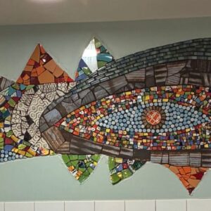 Mosaic mural with abstract eye shapes and colorful tiles decorates a wall above a row of white tiles; a light bulb hangs in front.