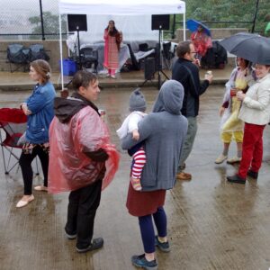 People wearing rain gear gather on a wet pavement under umbrellas near a canopy, as a person in front of a microphone stands under the canopy in the background.