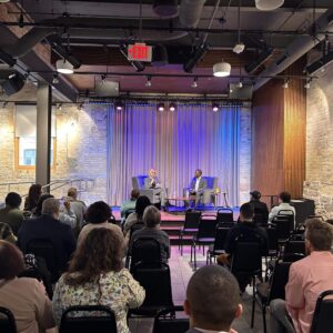 Two people sit on stage having a discussion in front of an audience in a modern event space with exposed brick walls and ceiling fixtures.