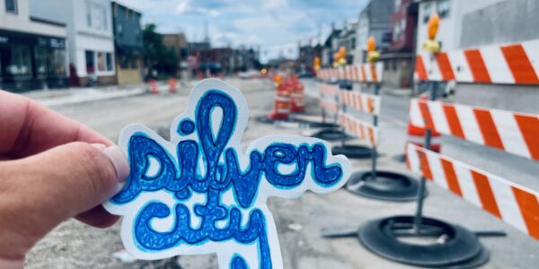 A hand holds a “silver city” sticker in front of a street under construction with orange and white barricades and houses on both sides.