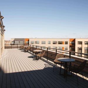 Outdoor terrace with wooden flooring, several round tables and chairs, and a view of modern apartment buildings in the background under a clear sky.