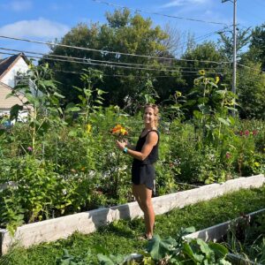 A woman stands in a sunny garden holding flowers, surrounded by tall plants and raised garden beds, with trees and power lines in the background.