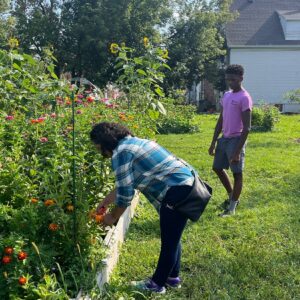 Two people are in a garden; one person is bending to pick or tend flowers in a raised bed while the other stands nearby on the grass.