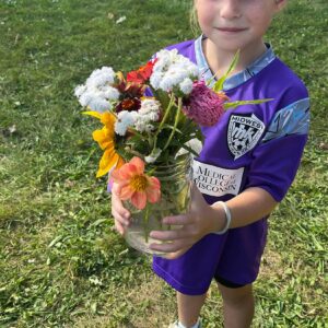 A young child in a purple soccer jersey holds a glass jar filled with assorted colorful flowers while standing on grass.