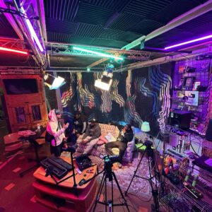 Four people sit on a couch in a studio with colorful lighting, cameras, audio equipment, and artwork on the wall, appearing to prepare for or record a podcast or interview.