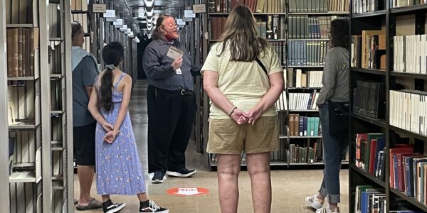 A group of people stands in a library aisle listening to a man speaking, surrounded by shelves filled with books.