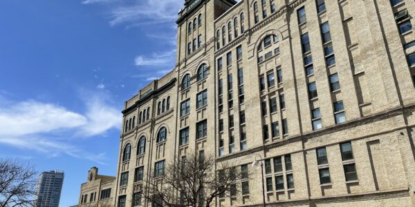 Large historic brick building with arched windows and a tower under a blue sky, surrounded by leafless trees and a tall modern building in the background.