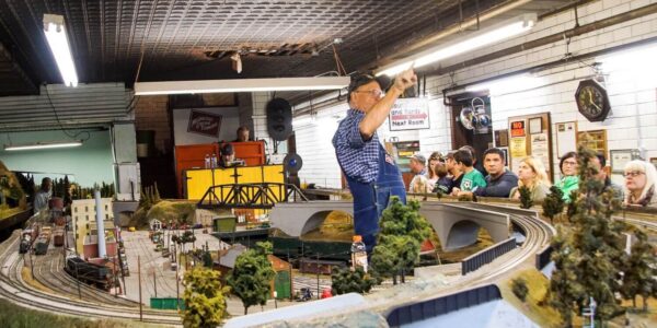 A man gestures over a large model train display, while a group of people watches from behind a glass partition in a room with industrial decor.