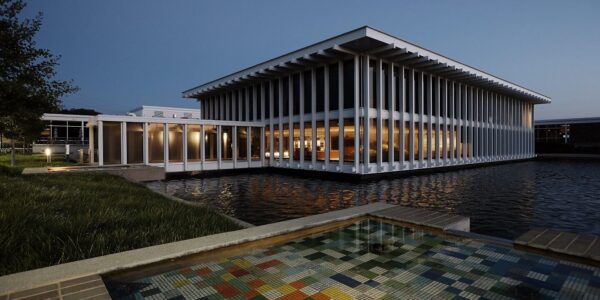 Modern building with large windows and vertical columns, surrounded by water. A tiled mosaic feature sits in the foreground, and the photo is taken at dusk.
