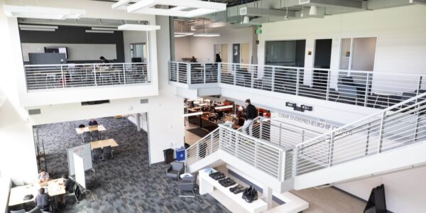 Spacious modern office interior with two floors, several people working at desks, large windows, and white railings surrounding an open central area.