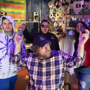 Four people pose in a colorful, tech-filled room. One person in front wears a plaid shirt and cap, holding up objects; three others stand behind, one in sunglasses, all holding small items.