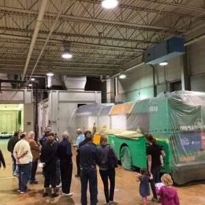 A group of people observes a large green bus covered in plastic inside an industrial facility with a high ceiling and bright lighting.