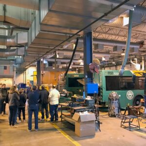 A group of people stands in a large industrial workshop with machinery, workbenches, and two green and blue buses inside.