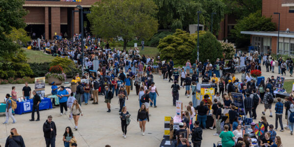 Large crowd of people walking among booths and tables at an outdoor campus event near brick buildings and greenery on a cloudy day.
