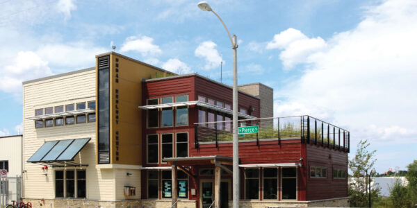 A modern, two-story building with large windows and a rooftop patio, located at the corner of a street with parked bicycles and a clear blue sky above.