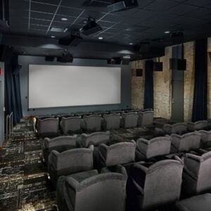 Empty theater room with several rows of gray cushioned seats facing a large blank screen, brick walls, and ceiling speakers.