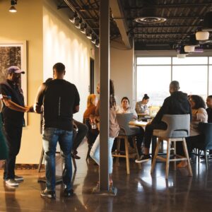 People are gathered in a modern, sunlit room, some standing and talking while others sit around tables, engaged in conversation.