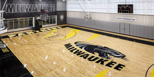 Indoor basketball court with “Milwaukee” and a panther logo on the floor, bright lighting, empty bleachers, and digital scoreboards on the back wall.