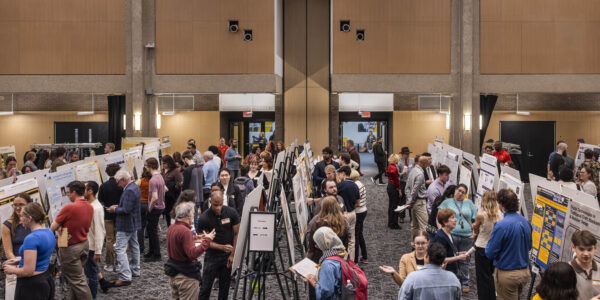 A large group of people view and discuss poster presentations at an indoor academic event in a spacious, well-lit hall.