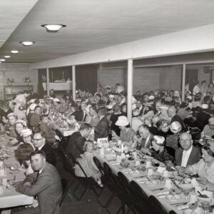 Large group of people seated at long tables indoors, dressed formally, eating a meal together in a crowded banquet hall.