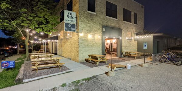 Outdoor seating area with picnic tables and string lights outside a brick building at night; several bicycles are parked near the entrance.
