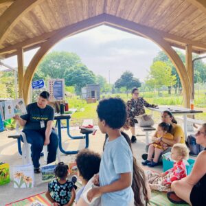 A group of adults and children sit under a wooden pavilion as a woman reads a picture book aloud; books are displayed on the ground nearby.