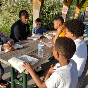 A group of children and an adult sit at a picnic table outdoors, reading and discussing books together on a sunny day.