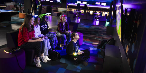 Five people sit and play video games together in a dimly lit arcade, focused on a large screen.
