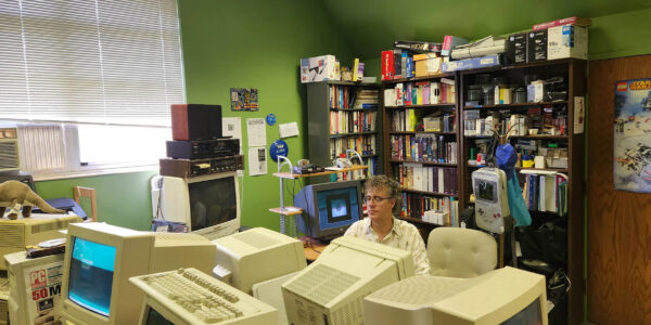 A person sits at a desk surrounded by vintage computer monitors in a cluttered office with green walls, bookshelves, and various electronics.