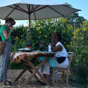 Two women interact at an outdoor table under a large umbrella, surrounded by sunflowers and greenery on a sunny day.