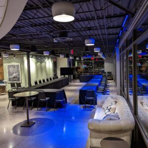 Modern event space with U-shaped tables, chairs, a couch, barstools, and large windows overlooking city lights at night. Ceiling lights and blue accent lighting illuminate the room.