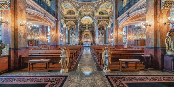 Ornate church interior with marble columns, wooden pews, statues, chandeliers, and detailed painted ceilings, viewed from the central aisle toward the altar.