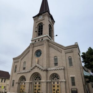 A beige brick church with a tall clock tower, arched windows and doors, a cross on top, and flower planters in front, photographed on a cloudy day.