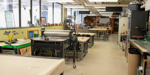 A spacious art studio with large windows, worktables, a printing press, and various art supplies and equipment arranged neatly around the room.
