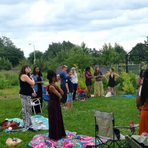 A group of people stands in a circle outdoors on a lawn, some with hands over their hearts, surrounded by blankets, chairs, and greenery under a cloudy sky.