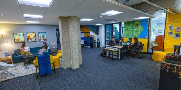 Office lounge with people sitting on couches and at tables, colorful decor, and maps and posters on the walls.