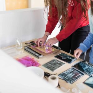 Two people work on an art project at a table covered with photographic prints, colored materials, and art supplies.