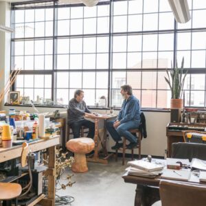Two people sit at a small table having a conversation in a bright, spacious studio filled with art supplies, tools, and large windows.