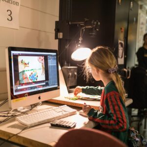 A young girl sits at a desk working on a stop-motion animation project on a computer, with art materials and a lamp nearby.