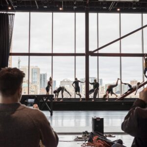 People rehearse a performance on a stage in front of large windows with a cityscape view, while two others observe and take photos from the audience area.