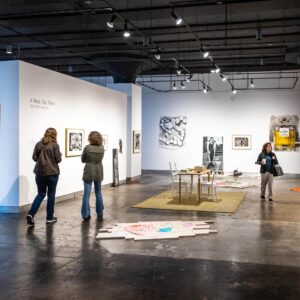 Three people view artwork displayed on white walls in a modern art gallery, with sculptures and framed pieces arranged throughout the brightly lit space.
