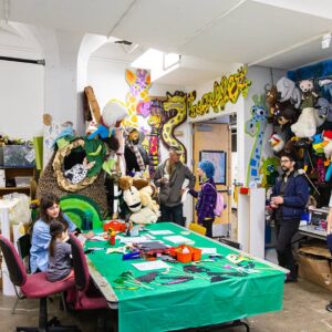 People gather in a colorful craft workshop filled with animal puppets, art supplies, and decorations on the walls. A green-covered table is at the center with craft materials.
