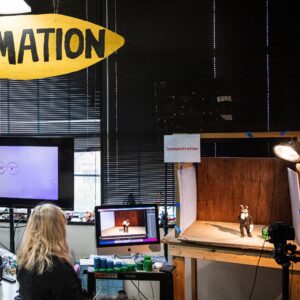 A person works at a stop-motion animation station, with computer screens, cameras, and a small model on a set under studio lighting. A large “ANIMATION” sign hangs above.
